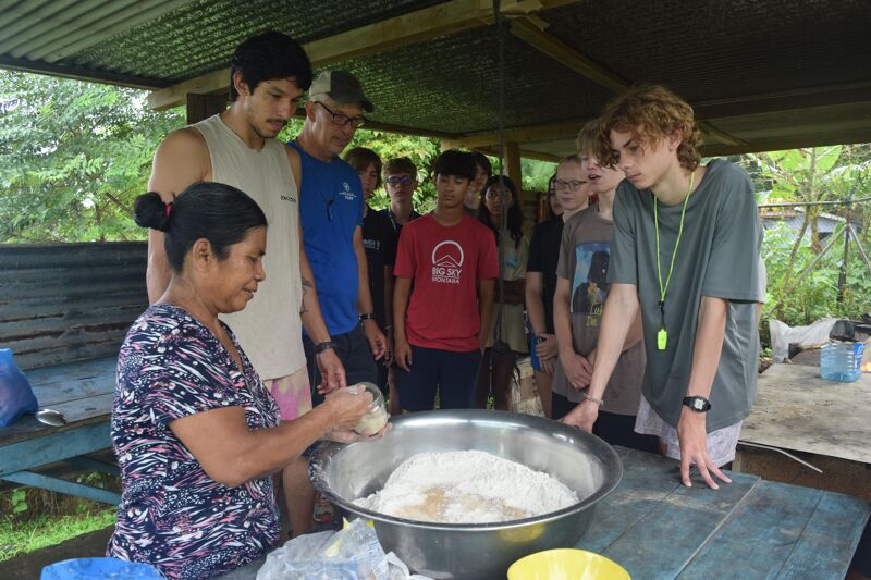 In the image, a group of people is gathered around a woman who appears to be demonstrating how to make something, possibly bread, using a large metal bowl. The people are of various ages and are looking at the bowl. The setting seems to be outdoors, under a covered area.